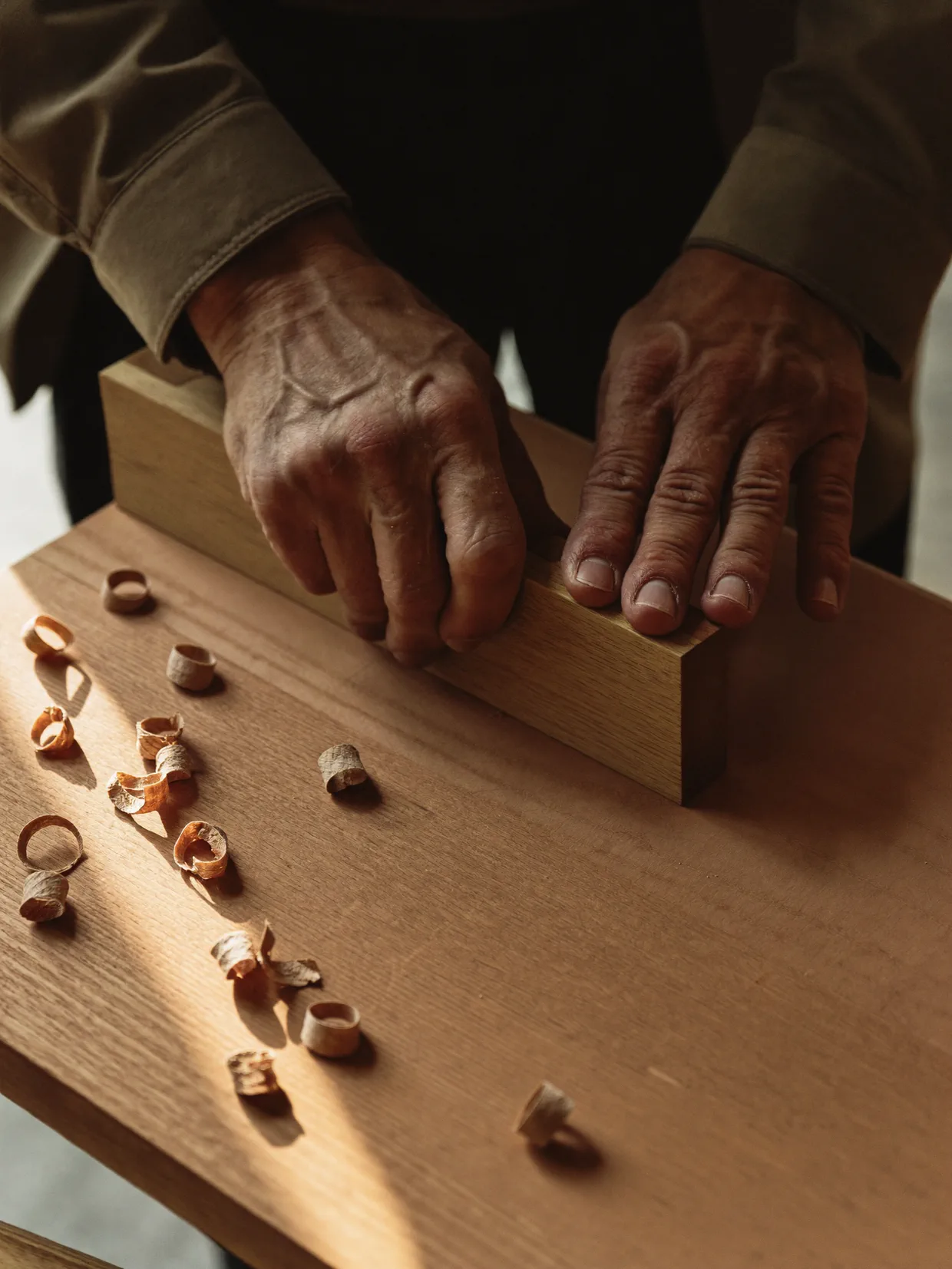 A pair of hands steadying a length of timber on a workbench, close in on the work itself, with curled wood shavings scattered across the surface.
