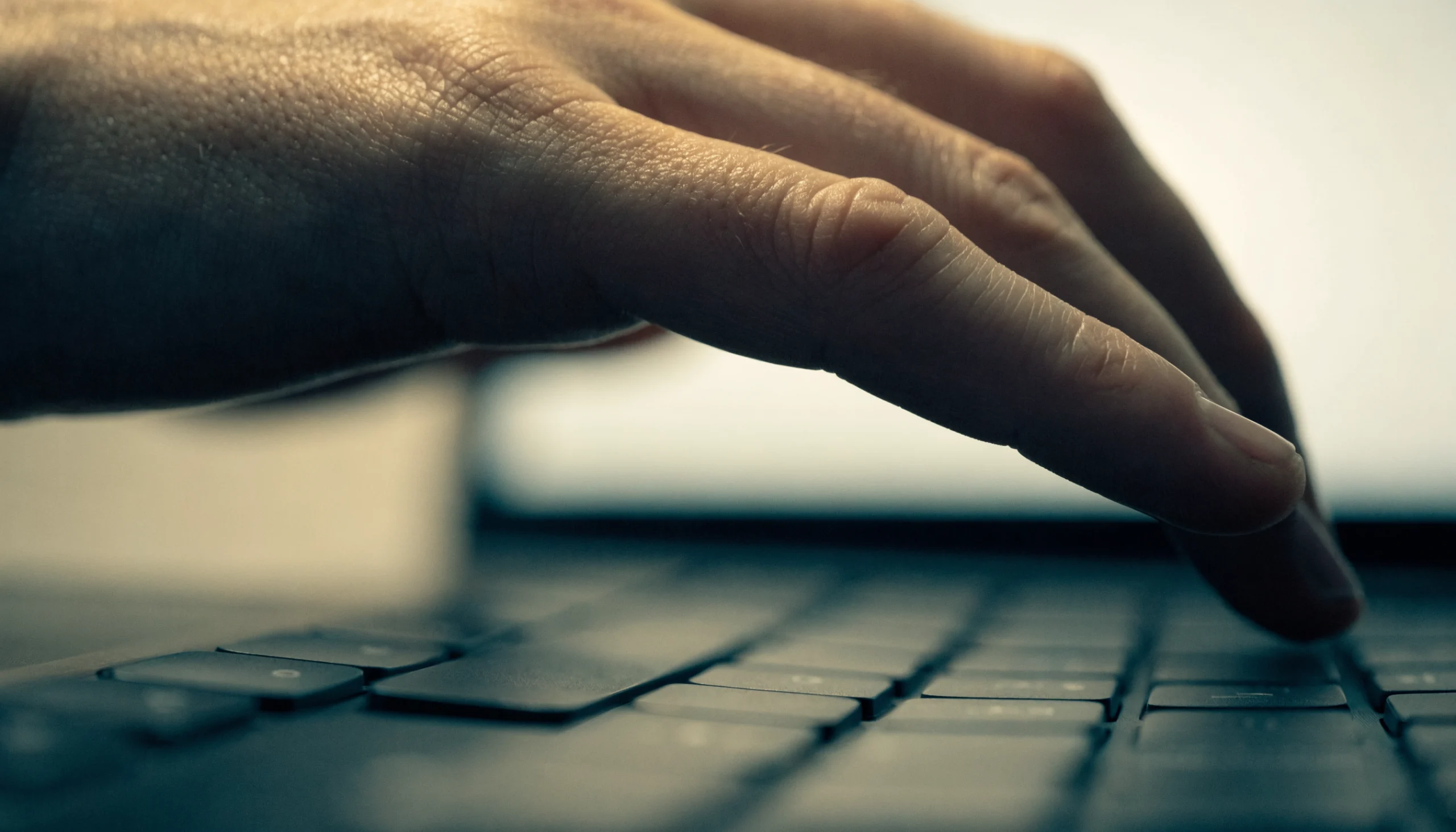 A hand held still just above a laptop keyboard, fingers not touching the keys, with the screen behind it out of focus